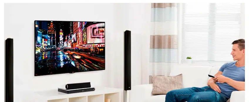 Hombre sentado en el sofá con el control remoto frente a un televisor colgado en la pared entre dos parlantes