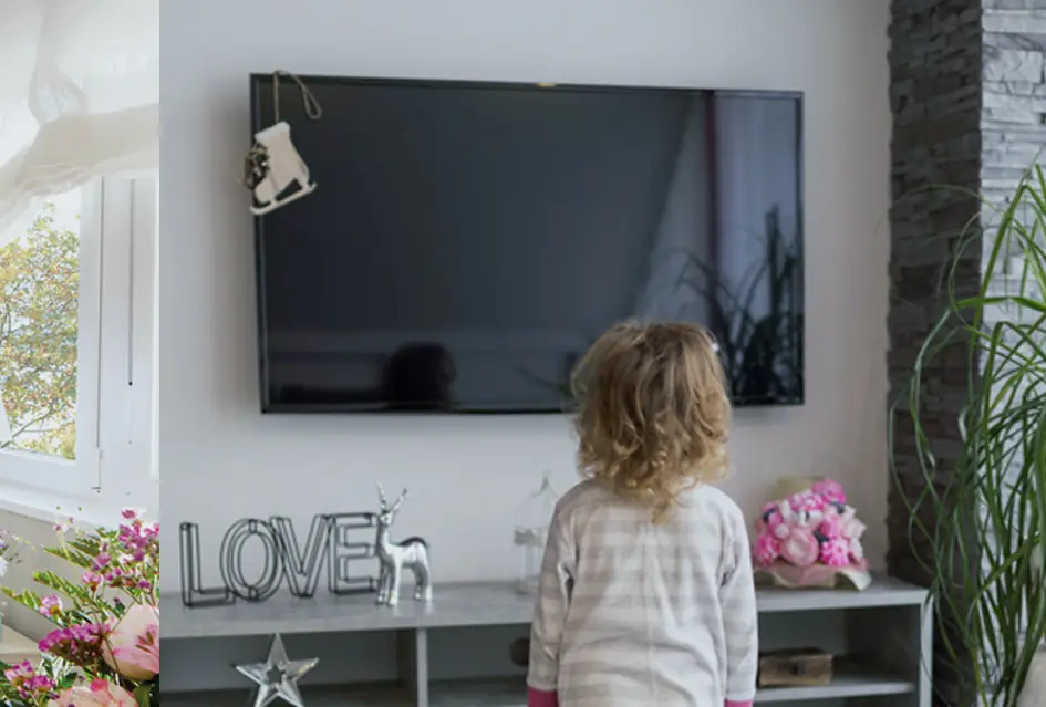 Niña de pie frente a un televisor colgado en la pared, sobre un mueble bajo